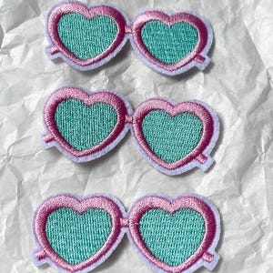 May include: Three embroidered patches in the shape of heart-shaped sunglasses. The frames are pink with a white border, and the lenses are a teal color. The patches are on a white, textured surface.
