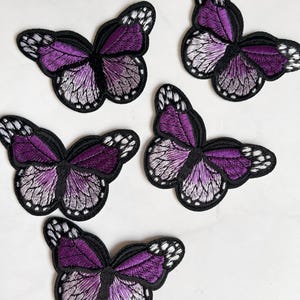 Può includere: Cinque toppe ricamate a forma di farfalla. Ogni farfalla presenta ali sfumate viola e bianche, bordate di nero con dettagli bianchi. Le toppe sono progettate per essere cucite o stirate sul tessuto.