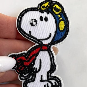 May include: Embroidered patch featuring Snoopy as a pilot, wearing a blue and yellow aviator helmet and a red scarf. The patch is white with black outlining and is a fun accessory for clothing or bags.