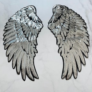 Puede incluir: Dos parches de alas de ángel con lentejuelas plateadas y bordes negros. Las alas son simétricas, con cada pluma detallada con pequeñas lentejuelas reflectantes. Estos parches son probablemente para la decoración de ropa o accesorios.