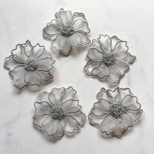 Könnte beinhalten: Fünf silber-graue Blumenapplikationen. Jede Blume hat ein zartes, durchscheinendes Blütenblattdesign mit silberner Kontur und einem strukturierten, grauen Zentrum. Die Applikationen sind auf einer weißen Oberfläche angeordnet.