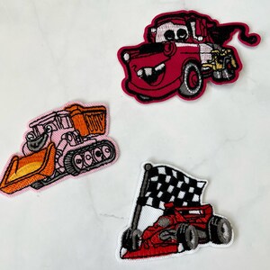 Pode incluir: Três patches bordados para passar a ferro que representam um camião de reboque vermelho, uma escavadora rosa e um carro de corrida vermelho com uma bandeira de xadrez.
