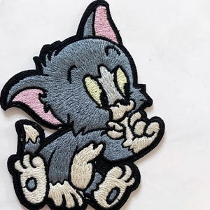 May include: Embroidered patch featuring a cartoon cat character in shades of gray, white, and pink. The cat is sitting with its paws in front, and has a black border. The patch is likely for clothing or accessories.