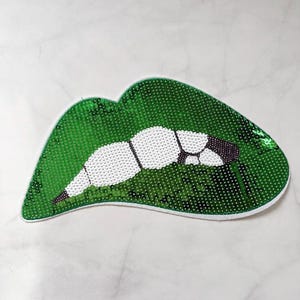 May include: A green sequined lips-shaped patch with white teeth and black accents. The patch is outlined in white and is on a white marble surface. The sequins give the patch a shiny appearance.