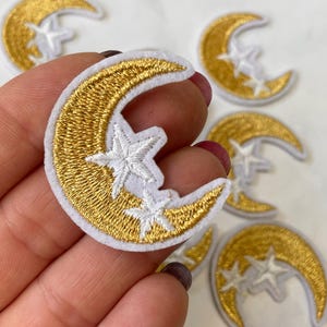 May include: A set of gold and white embroidered iron-on patches featuring a crescent moon with two stars.