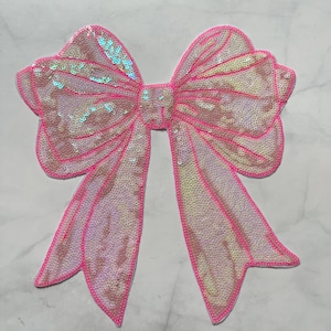 May include: A large, pink sequined bow with a bright pink outline. The bow has two long, pointed tails and is filled with iridescent sequins that catch the light. The bow is a decorative embellishment.