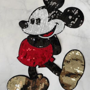 May include: A black and white sequined Mickey Mouse applique with red shorts and gold shoes.