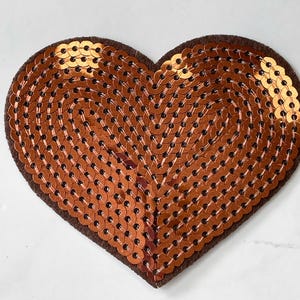 Puede incluir: Un parche en forma de corazón de color cobre, hecho de lentejuelas. Las lentejuelas están cosidas sobre un respaldo de fieltro marrón.