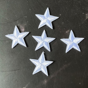 以下が含まれることがあります： 薄い青色の刺繍された星のパッチが5つ。各星には5つのポイントがあり、柔らかく質感のある生地で作られています。星は暗い背景に対して散らばったパターンで配置されています。