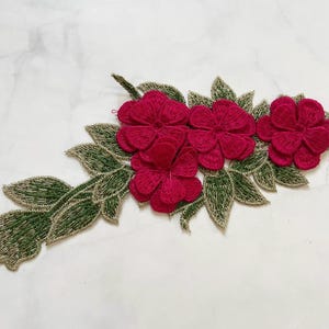 May include: Embroidered floral applique featuring five vibrant red flowers and green leaves with gold accents. This decorative embellishment is ideal for adding a touch of elegance to clothing, accessories, or craft projects.
