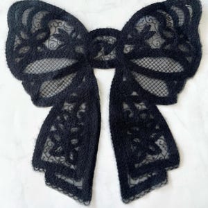 Può includere: Un fiocco di pizzo nero con un delicato motivo floreale. Il fiocco è realizzato in un tessuto morbido e trasparente e ha una texture leggermente pelosa.