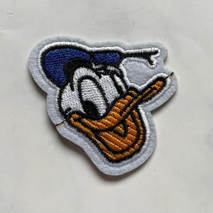 Puede incluir: Parche bordado con Donald Duck, un personaje de dibujos animados. El parche tiene un borde gris, con la cara de Donald en blanco, azul y naranja. El parche está diseñado para coser en ropa o accesorios.