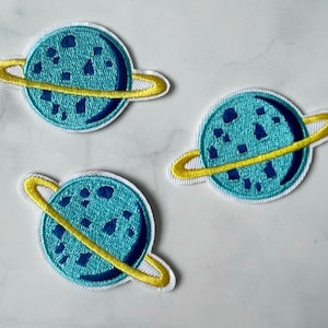 May include: Three blue and yellow embroidered iron-on patches of planets with rings. The planets are a light blue with dark blue spots and the rings are yellow.