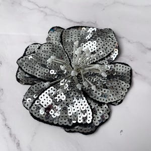 May include: A silver sequined flower applique with a pearl center. The flower has five petals and is surrounded by a black fabric border.