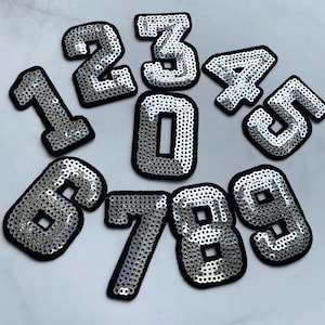 May include: Ten silver sequin iron-on patches in the shape of numbers 0 through 9. The numbers are outlined in black.