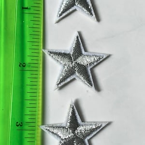 May include: Three silver star patches with white borders are arranged vertically. A green ruler with inch markings is on the left. Each star is approximately 2 inches in size, ideal for fabric embellishment or craft projects.