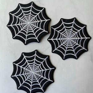 以下が含まれることがあります： 黒と白の蜘蛛の巣のパッチが3つ。パッチは黒い布地に白い糸で刺繍されています。