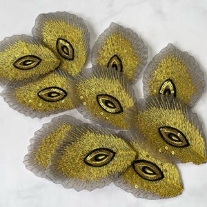 May include: A collection of gold embroidered leaf-shaped appliques. Each applique features a central eye-shaped design in black and gold, surrounded by gold thread and sequins. The edges are a sheer gray fabric.