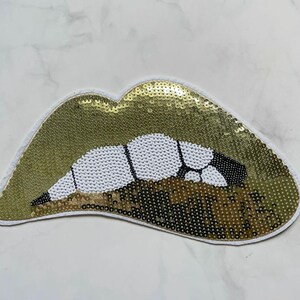 May include: A gold sequined lips patch with white and black detailing. The patch, in the shape of lips, has a white border. The sequins provide a shiny effect. This patch is suitable for clothing or accessories.
