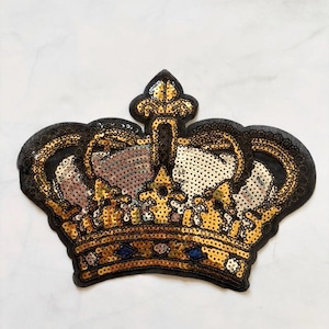 May include: A gold sequined crown patch with a black border. The crown features a detailed design with gold and silver sequins, and small blue accents. This decorative patch is suitable for embellishing clothing or accessories.