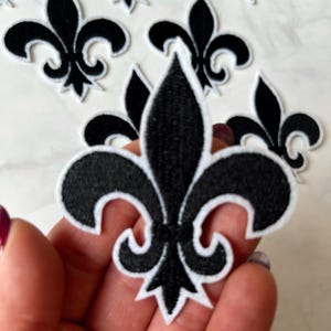 Pode incluir: Etiqueta termocolante preta e branca com um design de flor-de-lis. A etiqueta tem aproximadamente 5 cm de diâmetro.