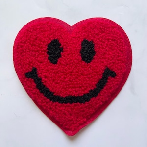 Puede incluir: Un parche rojo en forma de corazón con una cara sonriente negra bordada.