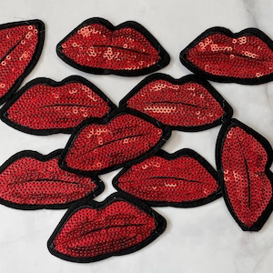 May include: Nine red sequin lips patches with black trim. The patches are arranged in a cluster on a white surface.