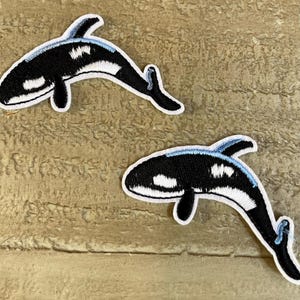May include: Two black and white embroidered orca whale patches with blue accents. The whales are facing opposite directions.