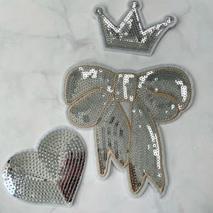 May include: Three silver sequin patches: a crown, a bow, and a heart. The patches are on a white background.
