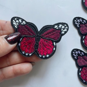Puede incluir: Parche de mariposa bordada con contorno negro, detalles blancos y una vibrante combinación de colores rojo y burdeos. El parche de mariposa está sostenido por una mano. Los parches son adecuados para ropa y accesorios.