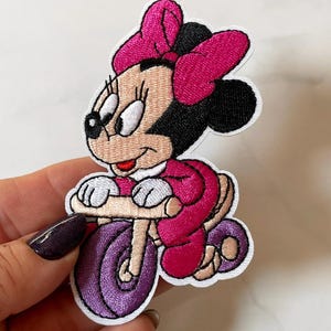 Pode incluir: Patch bordado com uma personagem de desenho animado num triciclo roxo. A personagem usa um laço e um vestido rosa, com rodas de triciclo a condizer. O patch tem uma borda branca, para roupa ou acessórios.