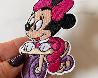 Könnte beinhalten: Gestickter Aufnäher mit einer Cartoonfigur auf einem lila Dreirad. Die Figur trägt eine rosa Schleife und ein Kleid mit passenden Dreiradreifen. Der Aufnäher hat einen weißen Rand, geeignet für Kleidung oder Accessoires.