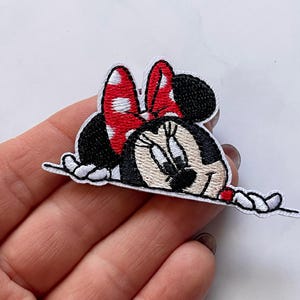 Peut inclure: Patch thermocollant brodé représentant un dessin animé de Minnie Mouse qui regarde par-dessus un fond blanc. Minnie porte son célèbre nœud rouge à pois blancs et a un visage noir et blanc.