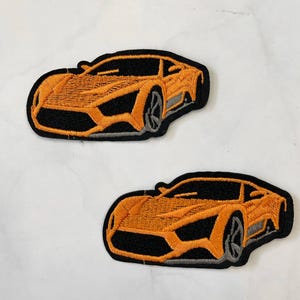 May include: Two orange embroidered iron-on patches of a sports car. The car is black with orange accents and gray wheels.