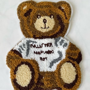 Könnte beinhalten: Ein brauner Teddybär-Plüsch-Patch mit einem weißen Shirt, auf dem "Maison Margiela" und "1977" in grauer Schrift stehen.