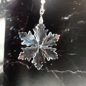 May include: A clear, faceted crystal snowflake ornament hanging from a silver-colored chain. The snowflake has six points with jagged edges, reflecting light. The background is a dark, marbled surface.