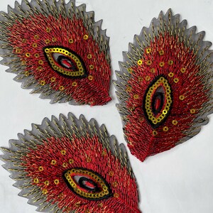 May include: Three red and gold peacock feather-shaped fabric appliques with sequins. The appliques are on a white background.