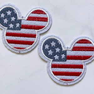Pode incluir: Dois patches em forma de Mickey Mouse com a bandeira americana em vermelho, branco e azul. Os patches são feitos de feltro e têm um contorno branco.