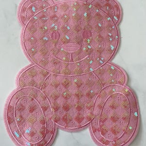 May include: Pink felt applique of a teddy bear with a checkered pattern of sequins. The bear has a simple embroidered face.