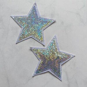 May include: Two star-shaped patches with a shimmering, iridescent surface. The stars are outlined with a white border. The patches are likely for decorative purposes, such as embellishing clothing or accessories.