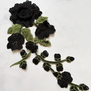 May include: Black lace floral applique with green leaves. The applique features a large rose and several smaller flowers, all in black. The leaves are a contrasting green color. This decorative applique is suitable for various craft projects.