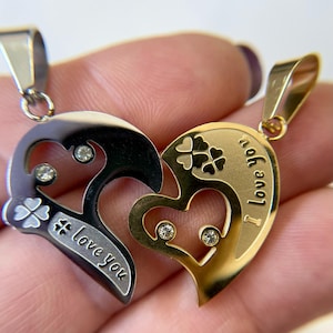 May include: Two heart-shaped pendants, one black and one gold, each with a portion of a heart design. The black pendant has the words "love you" and a clover. The gold pendant has the words "I love you" and clovers. Both have small, clear gemstones.