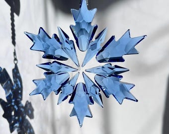Peut inclure: Ornement en forme de flocon de neige en cristal bleu, suspendu à une chaîne argentée. Les facettes du flocon projettent une ombre sur le fond blanc. Décoration sur le thème de l'hiver.