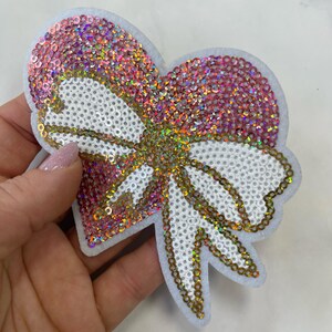 Può includere: Un cerotto a forma di cuore rosa e bianco con un grande fiocco bianco al centro. Il fiocco è bordato di paillettes dorate.