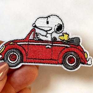 May include: Embroidered patch featuring Snoopy driving a red convertible. The patch has a white border and includes details like the car's wheels, headlights, and Snoopy's black ears. A yellow bird sits in the back seat.