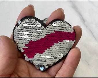 Puede incluir: Un parche de lentejuelas en forma de corazón de color plateado y rosa. Las lentejuelas están dispuestas en un patrón que crea una forma de corazón. El parche es perfecto para agregar un toque de brillo a cualquier proyecto.