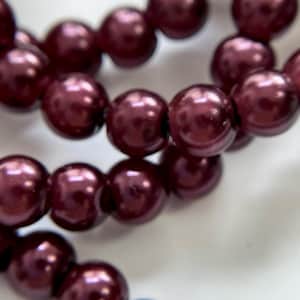 Op de afbeelding: Close-up van een streng bordeauxrode kralen. De kralen zijn rond en glanzend en reflecteren licht. De kralen zijn aan elkaar geregen, mogelijk een ketting of armband.