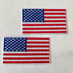 以下が含まれることがあります： アメリカの旗の刺繍パッチが2つ。旗は赤、白、青で、青いフィールドに白い星があります。パッチは白い背景にあります。