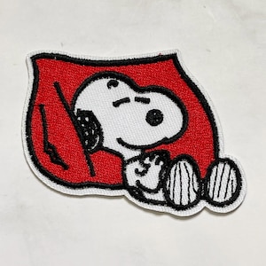May include: Embroidered patch featuring Snoopy from Peanuts, sleeping peacefully. The patch has a white Snoopy with black details, set against a red background with a black border. A charming accessory for clothing or bags.