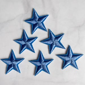 May include: Six blue embroidered star patches arranged in a triangular pattern. Each star has a lighter blue center and a darker blue outline, creating a dimensional effect. The patches are likely for decorative purposes.